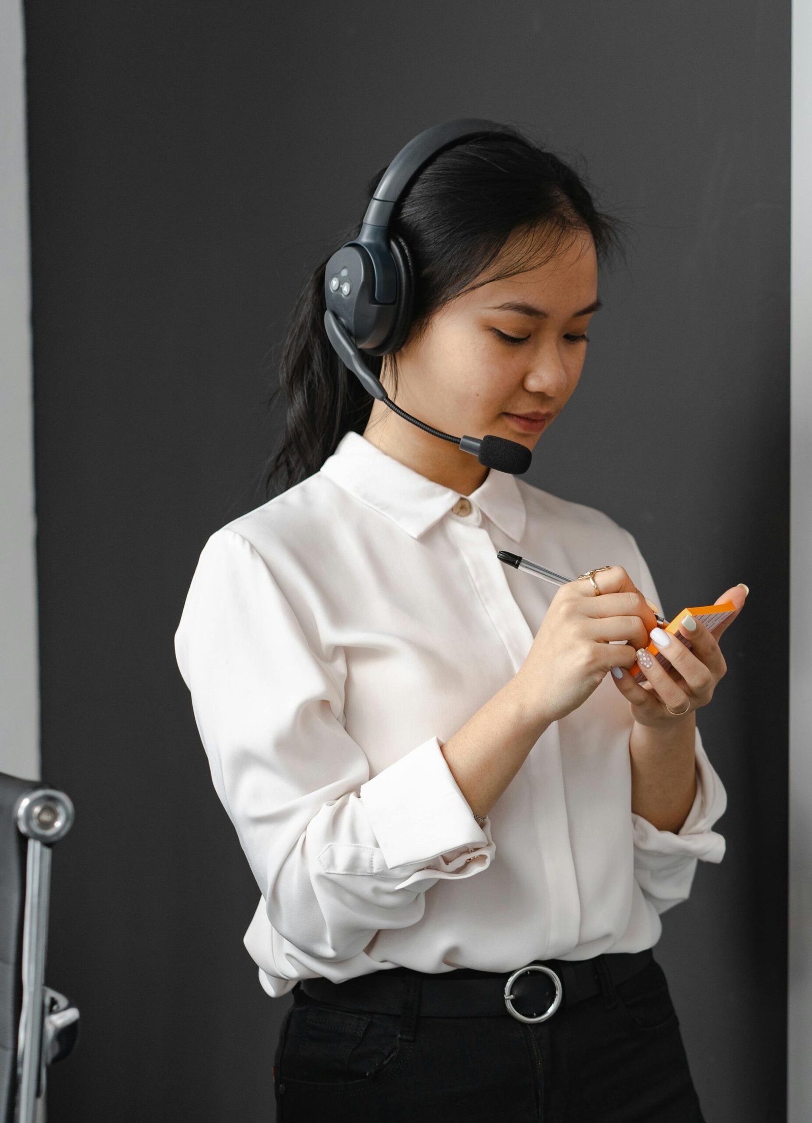 Asian woman in a white shirt with a headset writing notes, representing customer service.